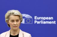 Mulher loira em blazer claro posa diante de fundo azul com logo e nome "European Parliament".