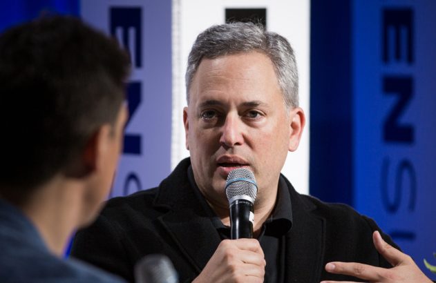 Homem de cabelo grisalho falando em um microfone, com outra pessoa de costas em primeiro plano à esquerda.