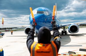 Pessoa em colete laranja guia avião azul da Southwest com bastões luminosos no pátio do aeroporto.