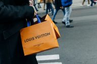 Sacolas Louis Vuitton laranjas com alças azuis nas mãos de uma pessoa em uma rua.