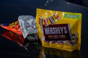 Três pacotes de chocolate: amarelo Hershey's Miniatures, prata Hershey's Kisses e laranja, em superfície reflexiva.