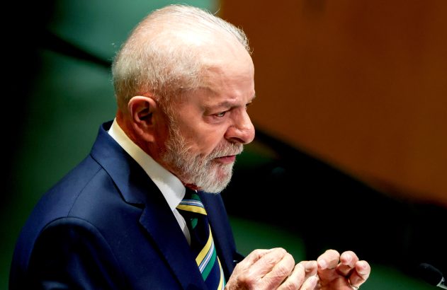 Lula, de terno azul e gravata verde e amarela, discursando em público com as mãos visíveis.