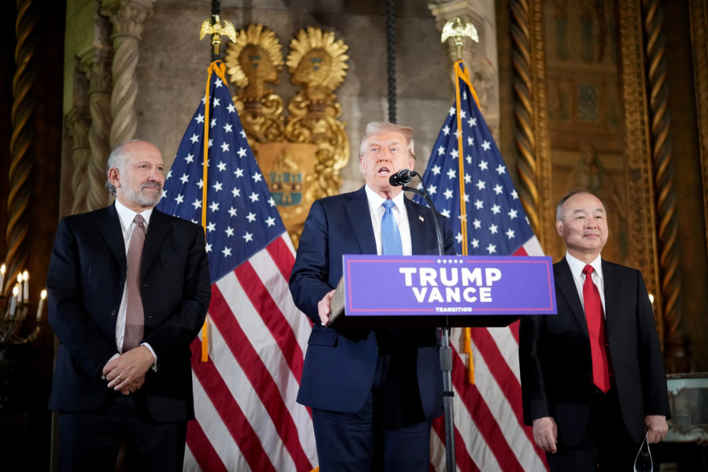 Donald Trump discursa em pódio, ladeado por dois homens. Bandeiras dos EUA e letreiro "TRUMP VANCE TRANSITION".