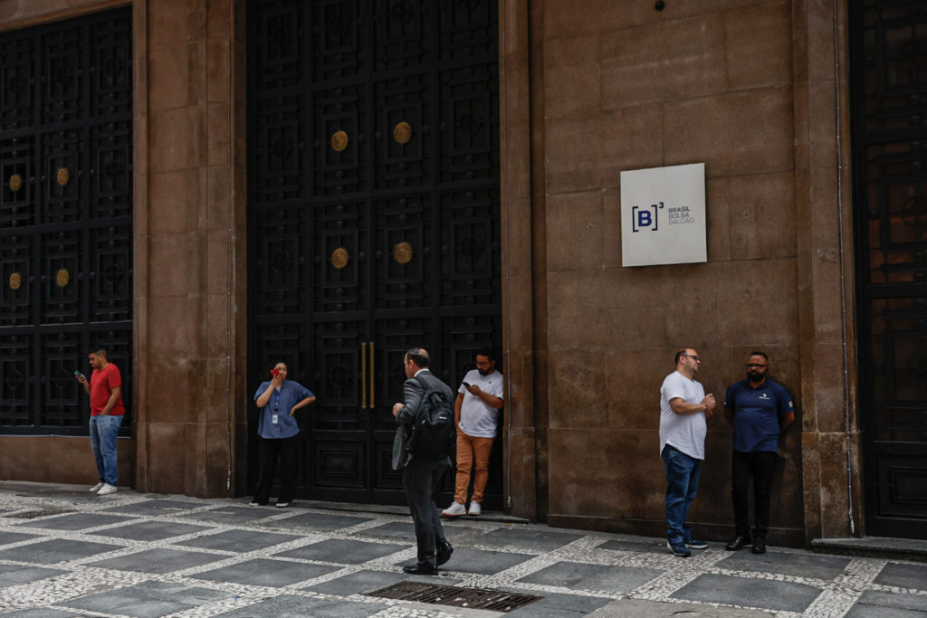 Grupo de pessoas em frente à B3 (Brasil Bolsa Balcão), alguns conversando, outros usando celular.