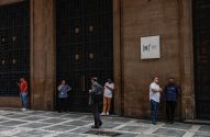 Grupo de pessoas em frente à B3 (Brasil Bolsa Balcão), alguns conversando, outros usando celular.