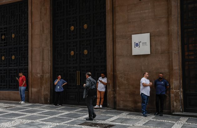 Grupo de pessoas em frente à B3 (Brasil Bolsa Balcão), alguns conversando, outros usando celular.