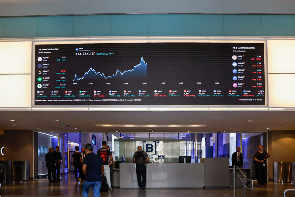 Telão digital exibe gráficos e dados de mercado em hall com pessoas passando.