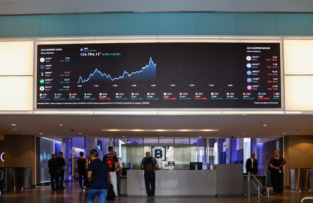 Telão digital exibe gráficos e dados de mercado em hall com pessoas passando.