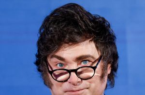 Close-up de homem de óculos e olhos azuis, com cabelo escuro e costeletas marcadas. Fundo azul liso.