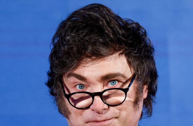 Close-up de homem de óculos e olhos azuis, com cabelo escuro e costeletas marcadas. Fundo azul liso.