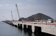 Longo cais de concreto em construção. Guindastes, materiais e van branca sobre ele. Montanha cinzenta com degraus ao fundo.