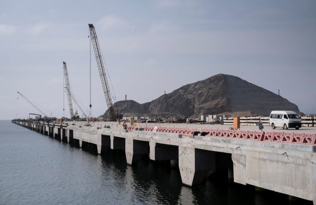 Longo cais de concreto em construção. Guindastes, materiais e van branca sobre ele. Montanha cinzenta com degraus ao fundo.