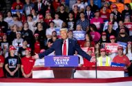 Donald Trump discursando em um comício, braços abertos, para apoiadores com banners políticos.