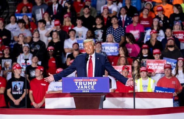 Donald Trump discursando em um comício, braços abertos, para apoiadores com banners políticos.