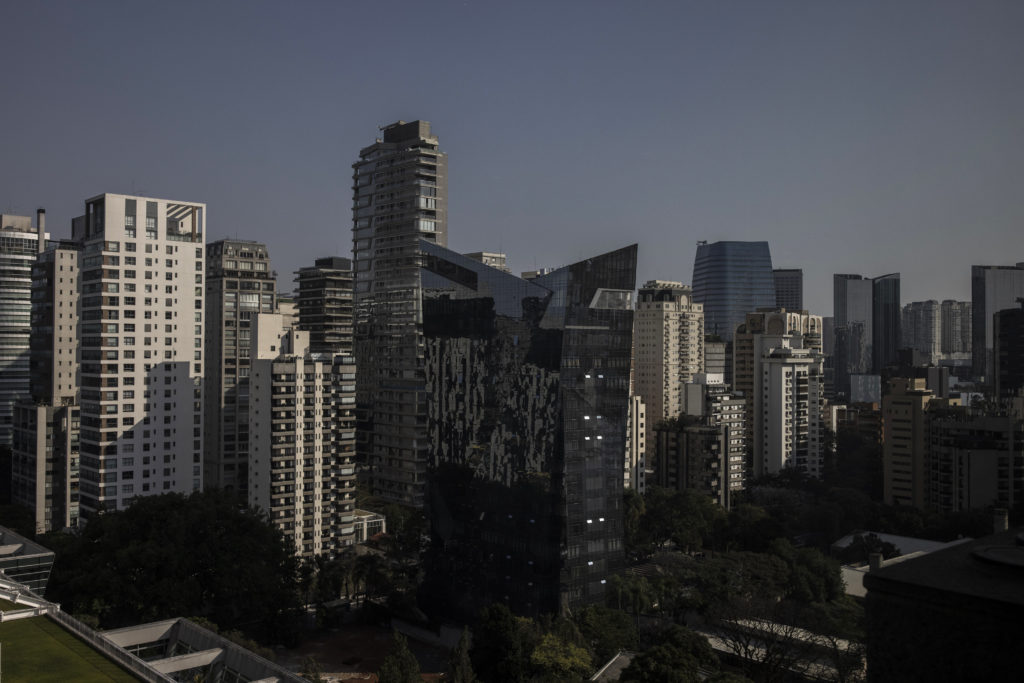 Vista panorâmica de uma cidade com diversos arranha-céus. Um prédio de vidro escuro e angulado se sobressai.