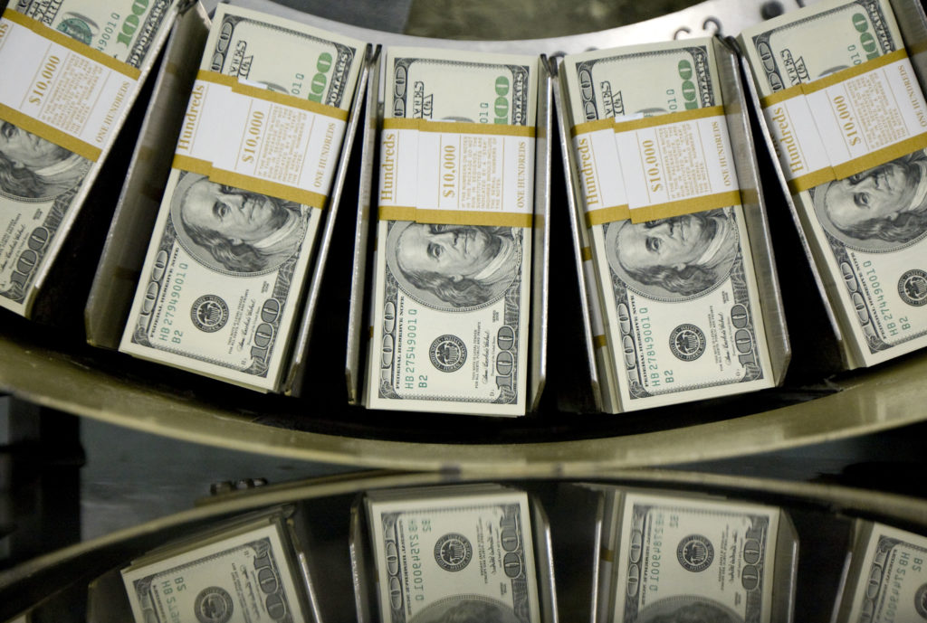 Maços de notas de US$100 em uma esteira de contagem, com reflexos visíveis.