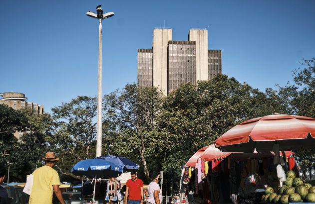 Pessoas em feira com barracas de guarda-chuva vermelho e azul, árvores e prédio no fundo, sob céu azul.