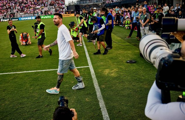 Lionel Messi, de branco, caminha por um campo de futebol, cercado por fotógrafos e outras pessoas.