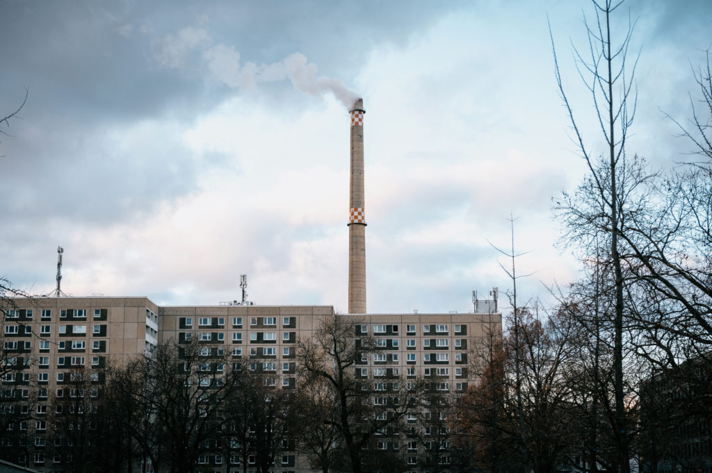 Chaminé industrial alta com fumaça, ladeada por edifícios e árvores nuas, sob céu nublado.