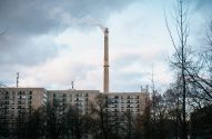 Chaminé industrial alta com fumaça, ladeada por edifícios e árvores nuas, sob céu nublado.