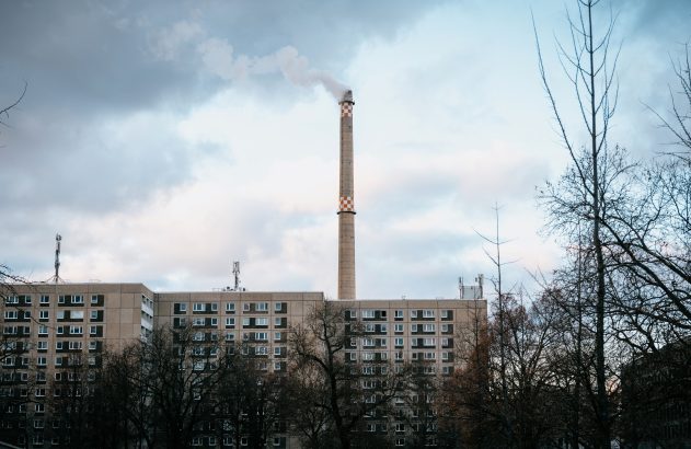 Chaminé industrial alta com fumaça, ladeada por edifícios e árvores nuas, sob céu nublado.
