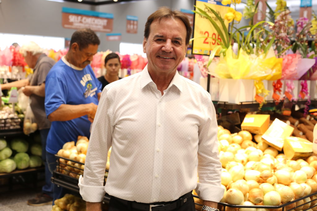 Homem sorridente em supermercado, seção de hortifrúti, com outros clientes ao fundo.