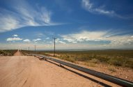 Estrada de terra, gasoduto e postes se estendem por paisagem árida sob céu azul e água ao fundo.