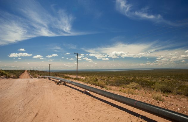Estrada de terra, gasoduto e postes se estendem por paisagem árida sob céu azul e água ao fundo.