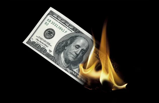 A imagem mostra uma nota de 100 dólares parcialmente consumida por chamas, destacando-se contra um fundo preto, simbolizando perda material.
