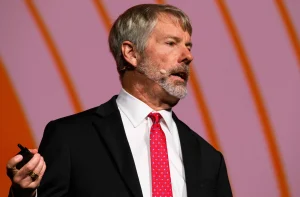 Michael Saylor é grisalho, com barba, está de terno preto e gravata branca e microfone
