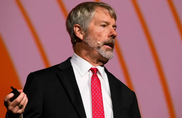 Michael Saylor é grisalho, com barba, está de terno preto e gravata branca e microfone