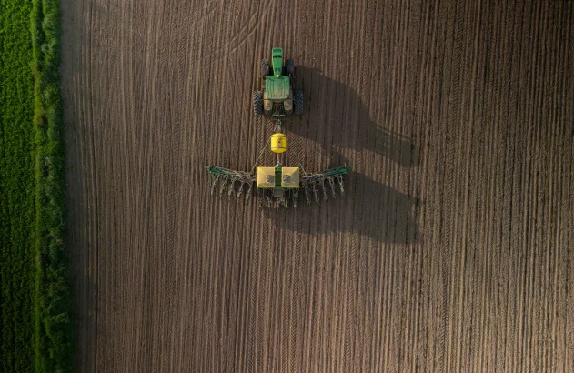 Imagem aérea de um trator verde com equipamento amarelo arando um campo, cercado por uma faixa de vegetação verde.