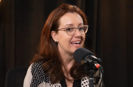 Mulher de óculos e cabelo ruivo sorri, falando em microfone preto em estúdio escuro.