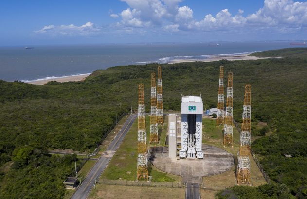 Complexo de lançamento brasileiro em área costeira. Torre com bandeira do Brasil, mastros e vegetação densa.