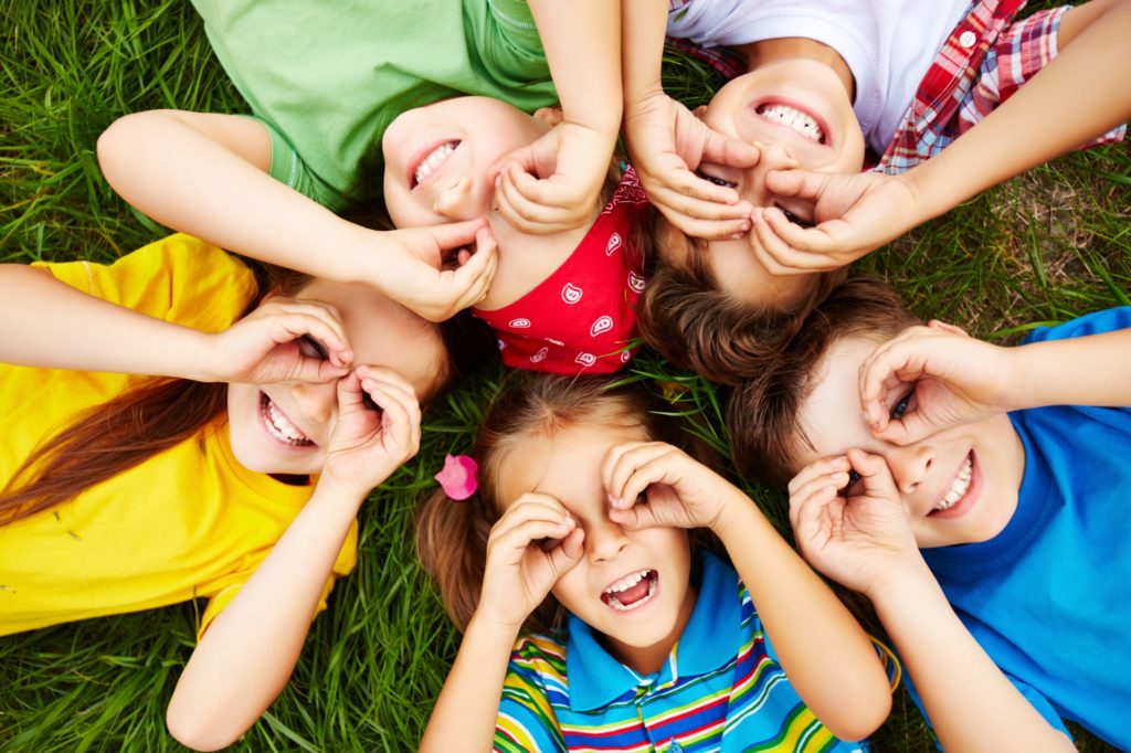 Crianças sorrindo e deitadas na grama, com as mãos formando binóculos nos olhos.