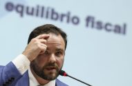 Homem barbudo de terno azul gesticula com a mão na testa. Microfone à frente, texto 'equilíbrio fiscal' desfocado ao fundo.