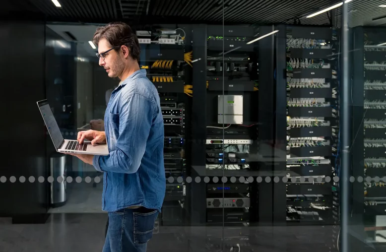 Um homem usando óculos e uma camisa jeans azul está concentrado em seu laptop em frente a um moderno datacenter com racks de servidores e cabos organizados.