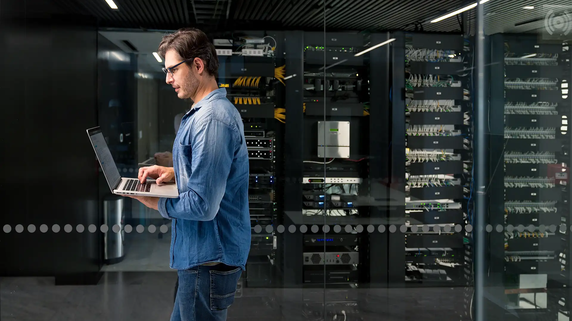 Um homem usando óculos e uma camisa jeans azul está concentrado em seu laptop em frente a um moderno datacenter com racks de servidores e cabos organizados.