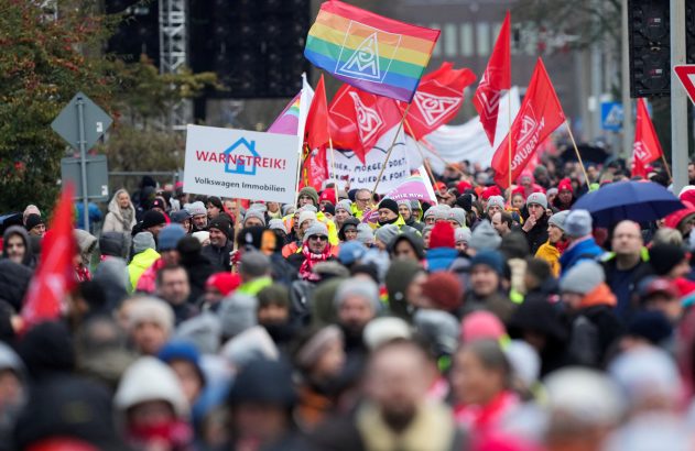 Multidão em greve, com bandeiras vermelhas e arco-íris, e um cartaz "WARNSTREIK! Volkswagen Immobilien".