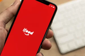 Uma mão segura um smartphone exibindo o aplicativo iFood em uma tela vermelha, com o logotipo branco em destaque e um teclado branco desfocado ao fundo.