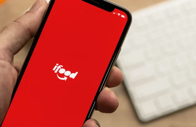 Uma mão segura um smartphone exibindo o aplicativo iFood em uma tela vermelha, com o logotipo branco em destaque e um teclado branco desfocado ao fundo.