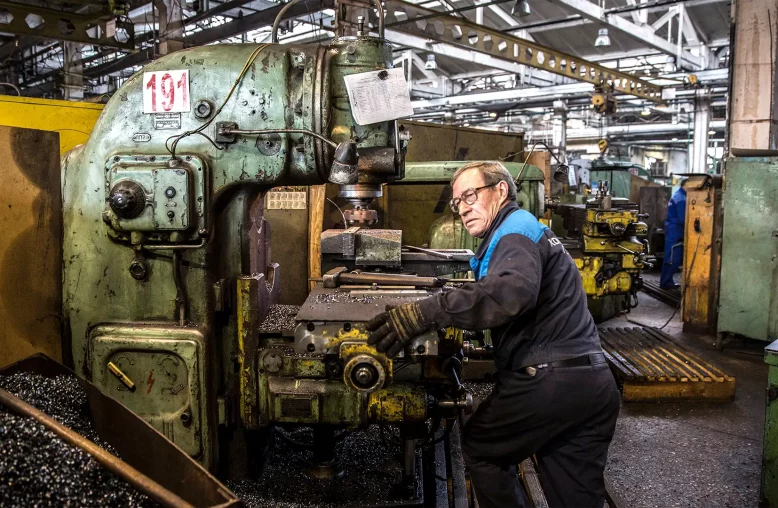 Um homem opera uma máquina industrial verde em uma oficina cheia de vida e atividade, com um ambiente de intensa usinagem.