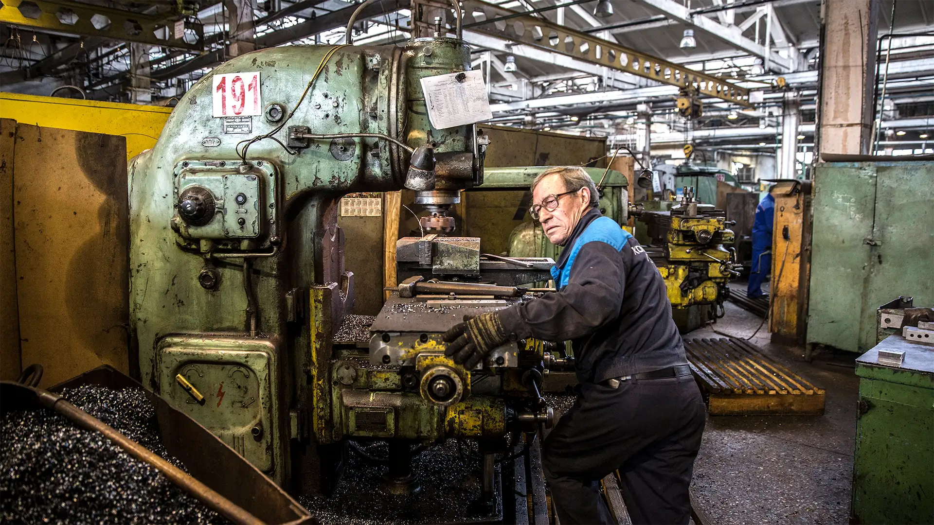 Um homem opera uma máquina industrial verde em uma oficina cheia de vida e atividade, com um ambiente de intensa usinagem.