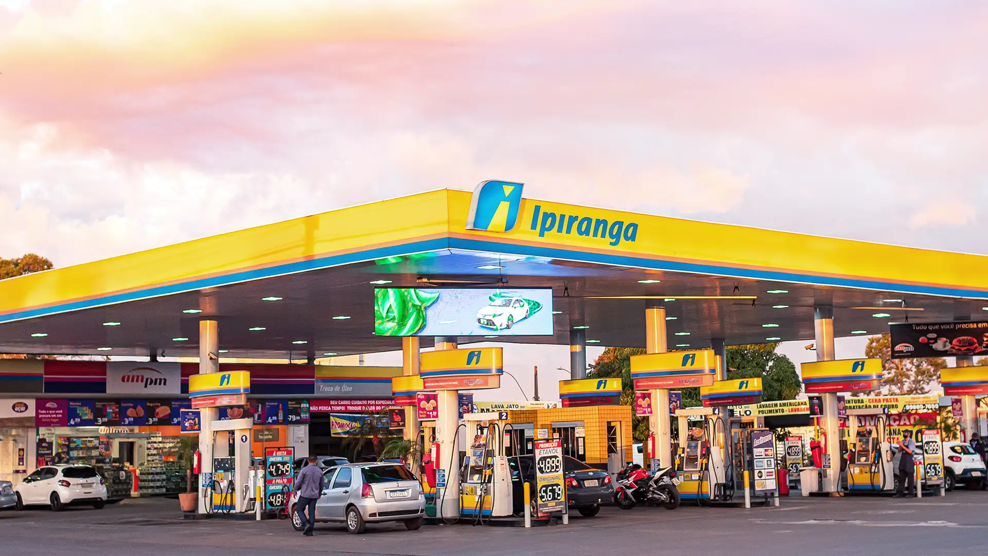 Posto de combustíveis da marca Ipiranga, com design moderno em amarelo e azul, bombas de abastecimento e uma loja de conveniência ampm, capturado ao entardecer com vegetação ao fundo.