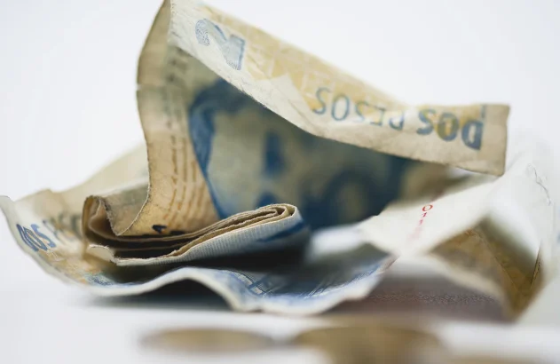 A imagem mostra cédulas amassadas de dois pesos argentinos sobre uma superfície clara, com foco na textura e nas dobras do papel.