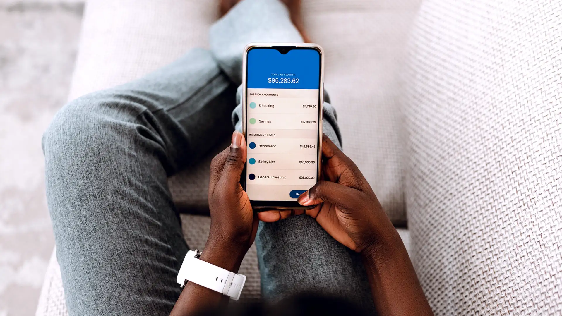 Uma pessoa deitada em um sofá cinza claro está usando um smartphone com um aplicativo financeiro aberto, mostrando seu patrimônio líquido total de $95,283.62.
