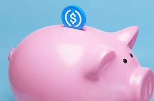 Cofrinho rosa brilhante com moeda azul sendo inserida no topo, sobre fundo azul claro.