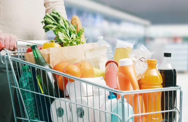 Carrinho de supermercado cheio com variedade de produtos, incluindo garrafas, sucos, papel higiênico, vegetais frescos, espaguete e laranjas, em um corredor de supermercado iluminado.