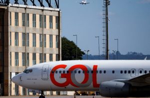 Avião GOL em solo, com outra aeronave se aproximando no céu. Ao fundo, um prédio e torre de iluminação de aeroporto.