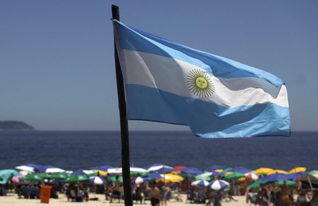 Bandeira da Argentina tremulando na praia com guarda-sóis e o oceano ao fundo.